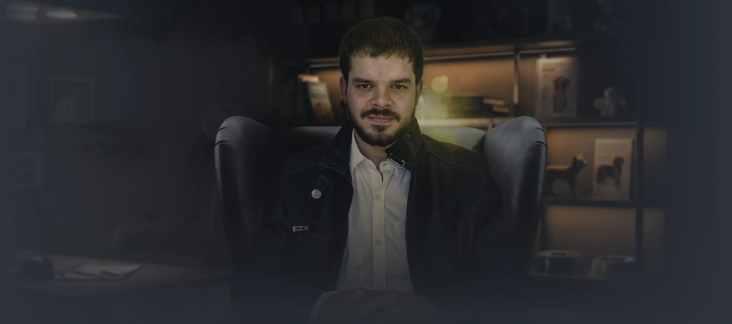 Um homem de barba e jaqueta de couro, sentado em uma poltrona em seu escritório veterinário